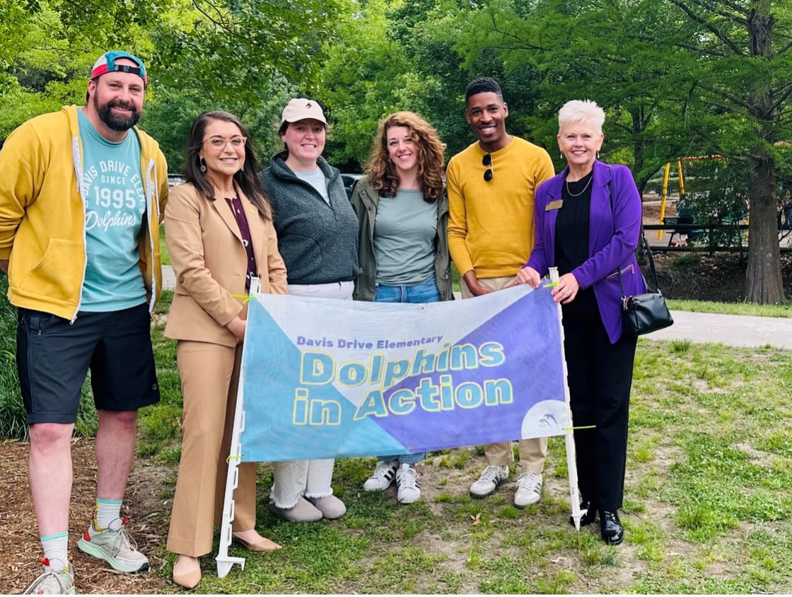 Six smiling people stand outdoors in a park holding a banner that reads "Davis Drive Elementary Dolphins in Action." The mood is cheerful and collaborative.