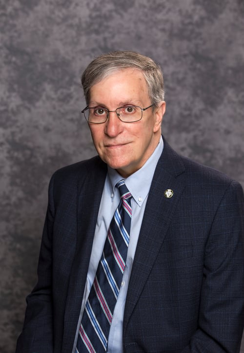 Image of an older man sitting in front of a gray photo backdrop. 