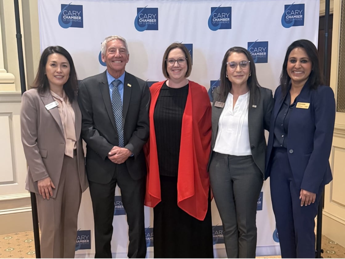 Five people stand smiling in front of a "Cary Chamber" backdrop. They are dressed in professional attire, conveying a formal and positive tone.