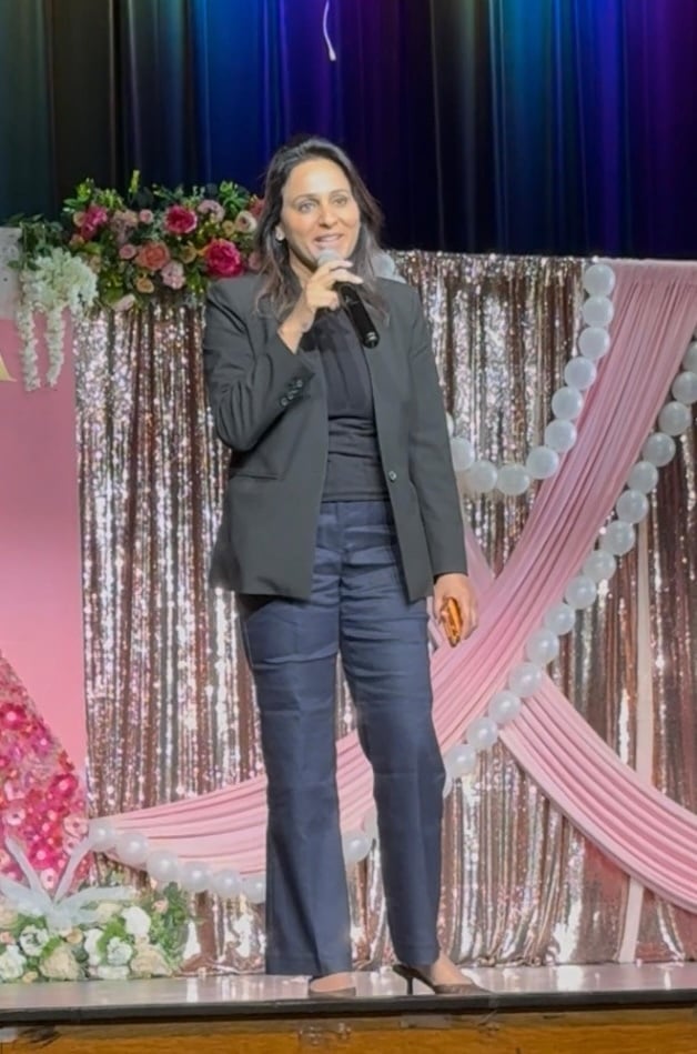 Woman in a black suit speaks onstage, holding a microphone. The backdrop features pink and white floral arrangements and sparkly curtains. 