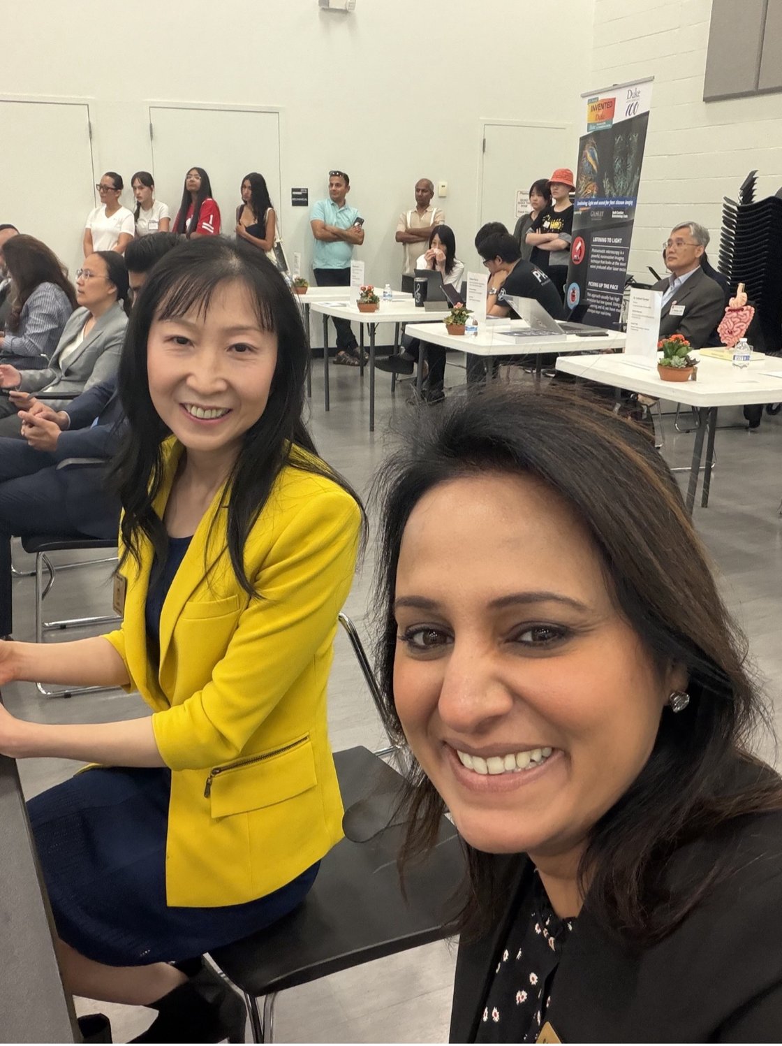 Two smiling women, one in a yellow jacket, take a selfie at a professional event. People stand and sit at tables in the background, creating a lively atmosphere.